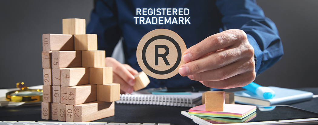 Person holding registered trademark symbol over desk, REGISTERED TRADEMARK text, with wooden blocks and office items.