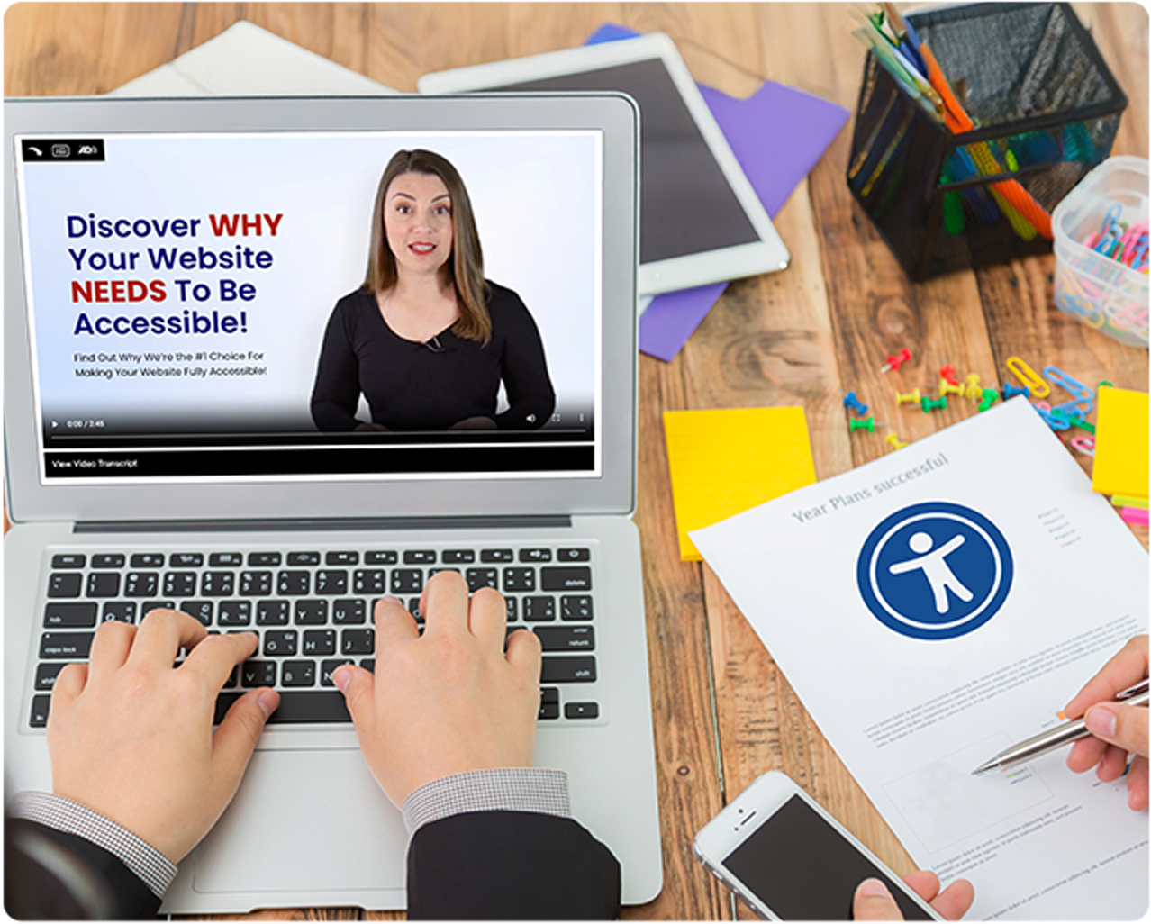 Person typing on a laptop with a video about website accessibility, surrounded by office supplies and a plan with an accessibility symbol.